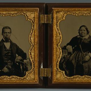 Duas fotografias em preto e branco em uma caixa de madeira com dobradiças e ouro. O homem, à esquerda, e a mulher, à direita, são negros e usam um vestido e terno escuro da moda.