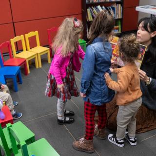 A woman with black hair shows a book to four toddlers in a room with colorful chairs.