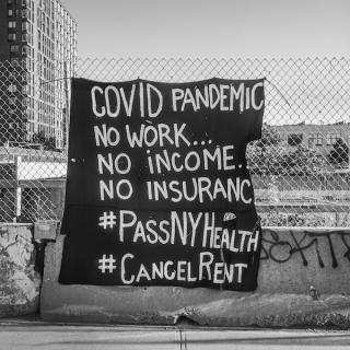 Uma fotografia em preto e branco de uma placa em uma cerca que diz "COVID PANDEMIC No work .... No Income ... No Insurance ... # PassNYHealth #CancelRent"