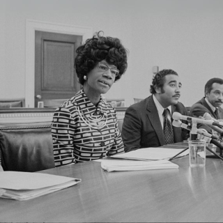 Uma mulher de óculos fala em uma mesa de conferência com dois homens ao lado dela e microfones na frente deles.