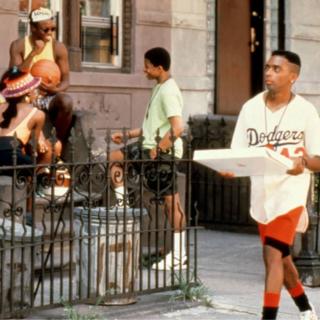 Spike Lee enquanto Mookie caminha por uma rua Bedford-Stuyvesant carregando uma pizza