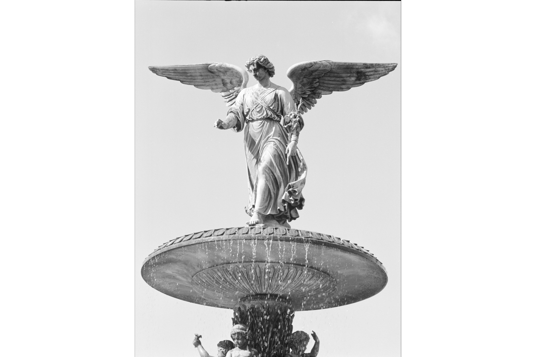 Bethesda Fountain: A black-and-white photograph shows a winged angel statue standing atop a large fountain, arms gently extended as water cascades below against a bright, open sky.