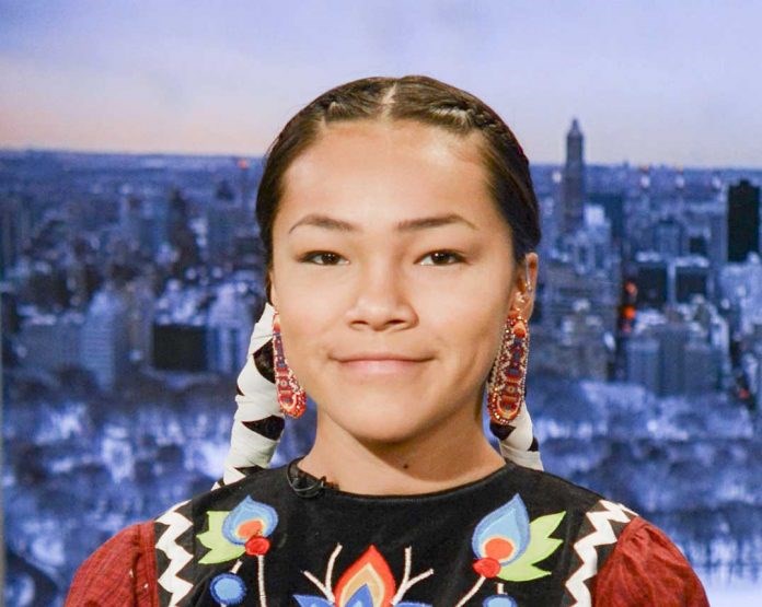 Autumn Peltier sits in front of a superimposed city skyline. She is wearing a garment with blue, red, green and yellow flowers embroidered on a black fabric which covers the front of a red checkered fabric. Peltier has two braids wrapped in beige ribbons, and she wears colorful beaded earrings. Peltier smiles directly at the camera.