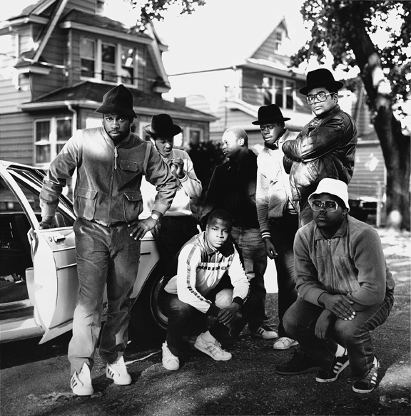 Fotografia em preto e branco com o Run DMC posando na frente de um carro em uma rua residencial.