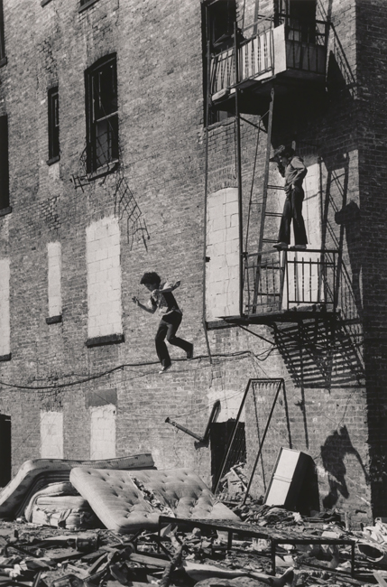 Black and white photograph of a brick building. A boy stands on the railing of a fire escape looking down at another boy who is captures mid-air before he lands on a pile of old mattress.