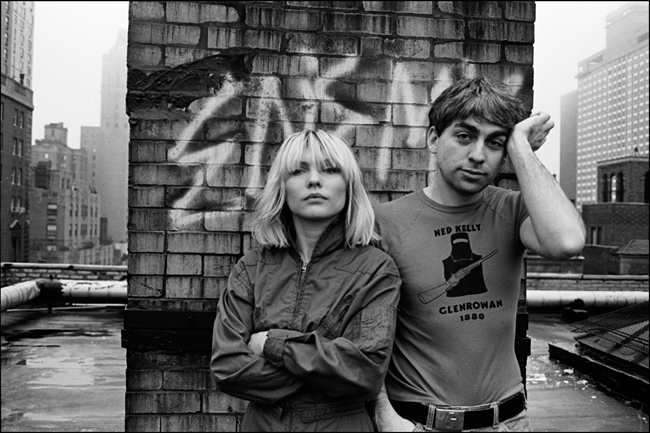 Black and White photograph of a man and woman standing on a roof, facing the camera. The woman has her arms crossed, the man holds a hand up to his temple.