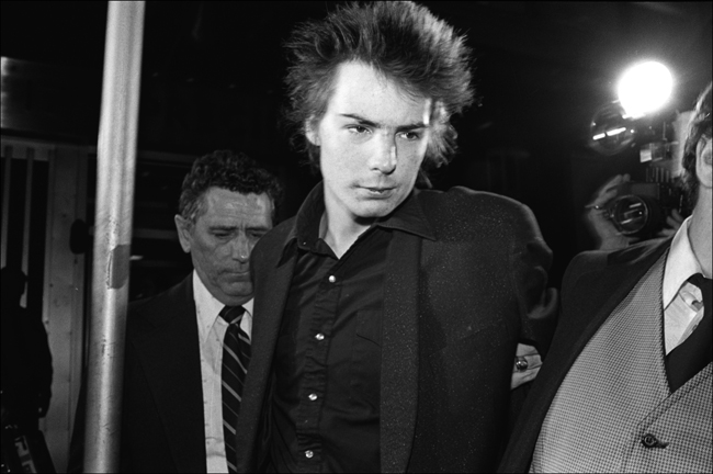 Photographie en noir et blanc de Sid Vicious. Un homme à sa gauche attrape son bras et l'emmène.