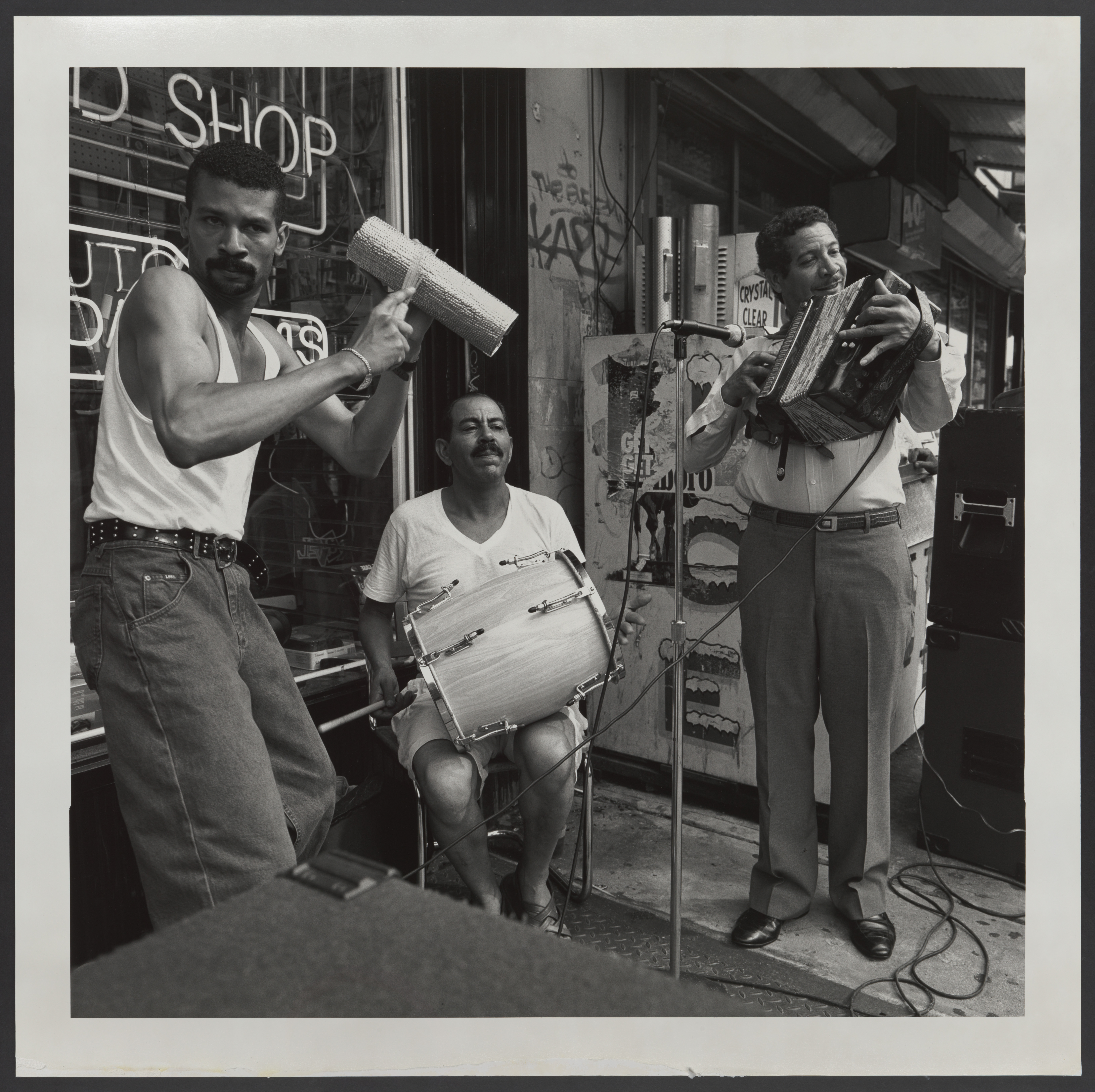 Black and white photograph of three men playing musical instruments outsde of a shop. One man is seated with a drum on his lap while the other two are standing, with one strumming a guira and another smiling as he plays his instrument.