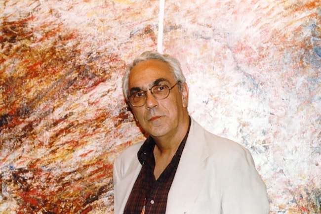 Color photograph of Mario César Romero wearing a white suit jacket and burgundy button down shirt against a brown-marbled wall