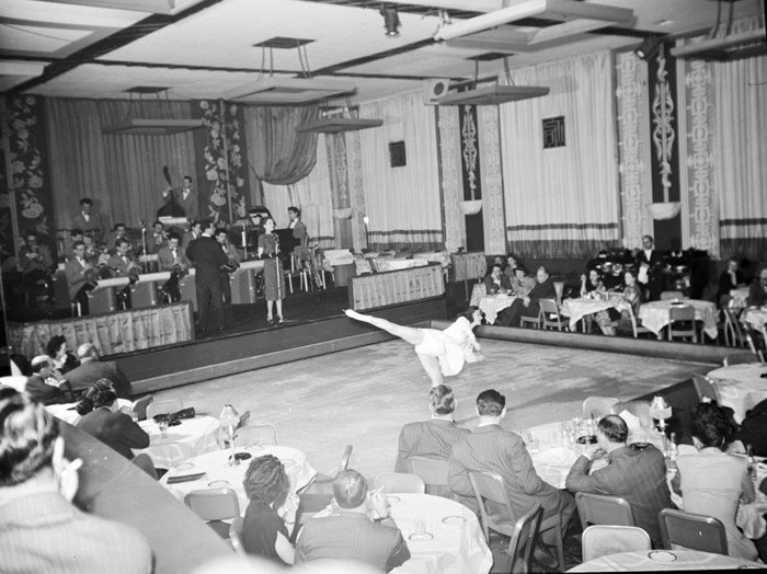 Skater Alice Farrar performs on an indoor ice rink, surrounded by people seated in tables and chairs at the Terrace Room, with a full band and singer in the background..