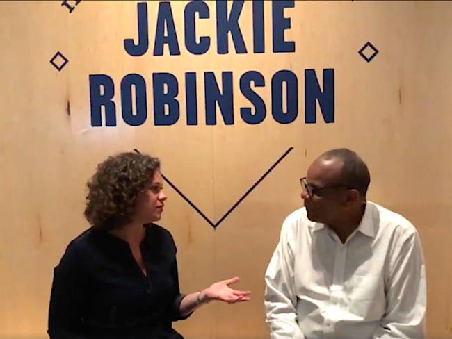 A man and woman sit in front of signage for the exhibition "In the Dugout with Jackie Robinson: An Intimate Portrait of a Baseball Legend."