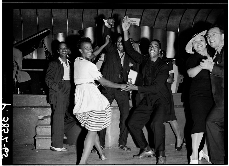 A lively group of Black men and women dance the Jitterburg and smile joyfully at nightclub in Harlem, 1939. A woman in a striped dress twirls with a partner at center, while others clap, cheer, and dance nearby. A jazz band is visible in the background, with a pianist and a saxophonist playing on stage. The atmosphere is energetic and celebratory.