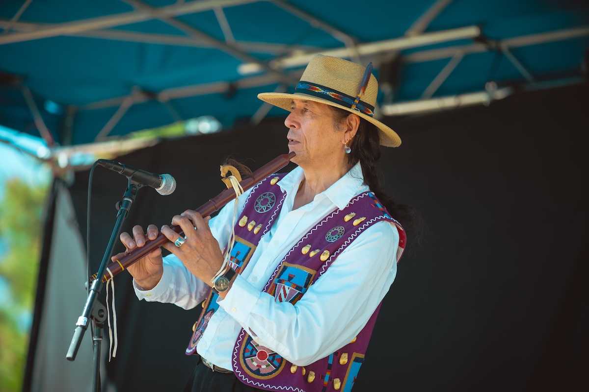 A man in a large hat and traditional style vest plays a flute. 