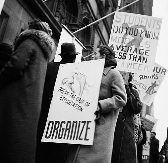 A fotografia mostra modelos da indústria de vestuário protestando contra os salários de trabalho da Art Students League. A imagem é em preto e branco e tem manifestantes segurando cartazes que dizem "Organize" e "Rompa o domínio da exploração"