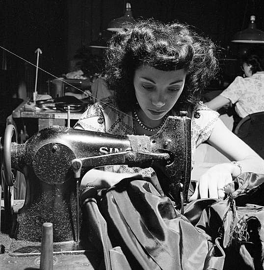 Photograph is black and white and shows a young woman working in a garment factory in 1949. The photograph was taken for Look magazine.