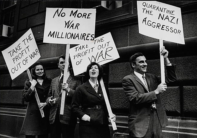 Black and white photograph depicts a group of men and women protesting American involvement in conflicts abroad. Protestors are holding signs addressing their concerns.