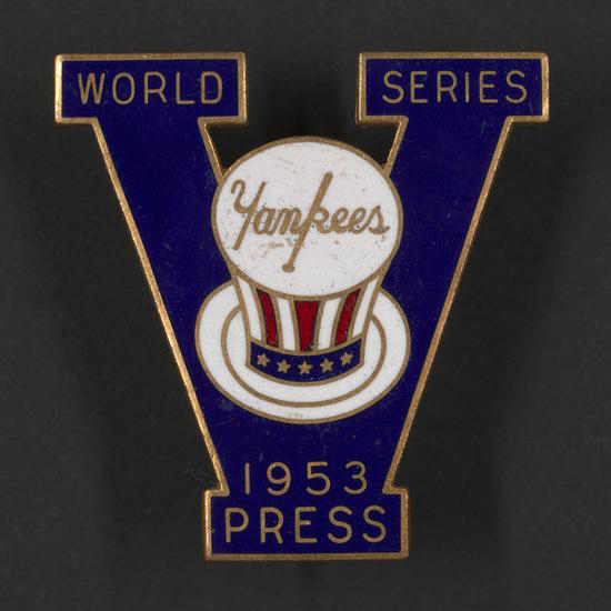 Enamel pin in the shape of the letter “V.” Top left of “V” says “World,” and top right says “Series.” At center is Yankees logo on stars and stripes top hat. At the bottom, “1953, Press.”