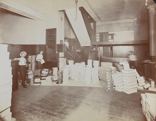 Interior del almacén de almacén de Rogers Peet & Co. con dos hombres trabajando y cajas apiladas alrededor.