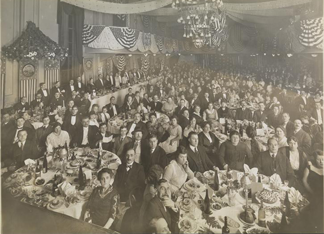 Fotografía en blanco y negro alrededor de 1910 de un banquete formal. Hombres, mujeres y algunos niños se sientan en las mesas mirando a la cámara, los cubiertos, los postres y las botellas de vino son visibles en las mesas.