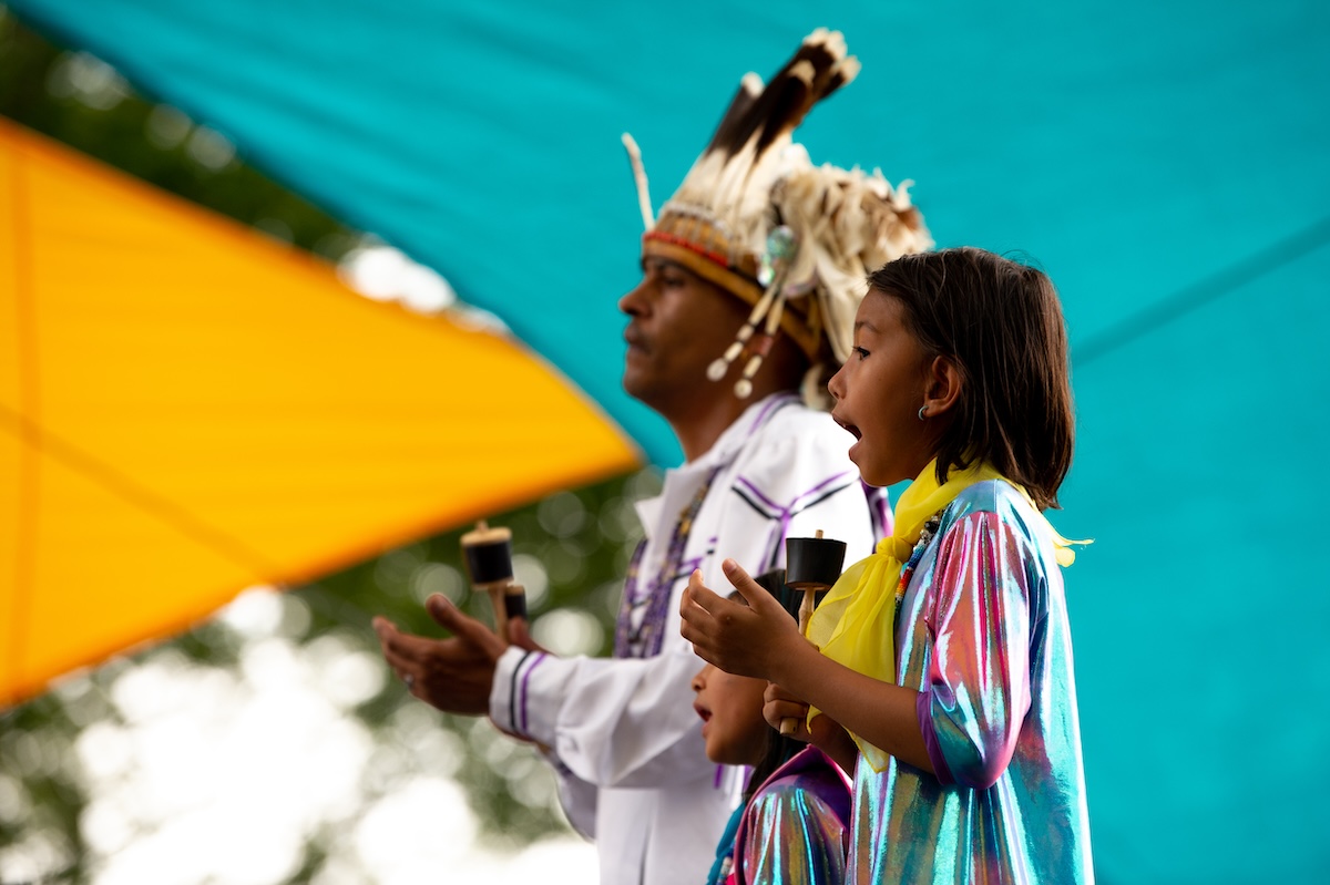 Um homem e uma jovem com roupas tradicionais cantam diante de um cenário turquesa.