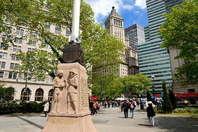 Photograph of people walking around a park surrounded by tall buildings in the background. A memorial statue with a plaque stands off-left in the foreground.