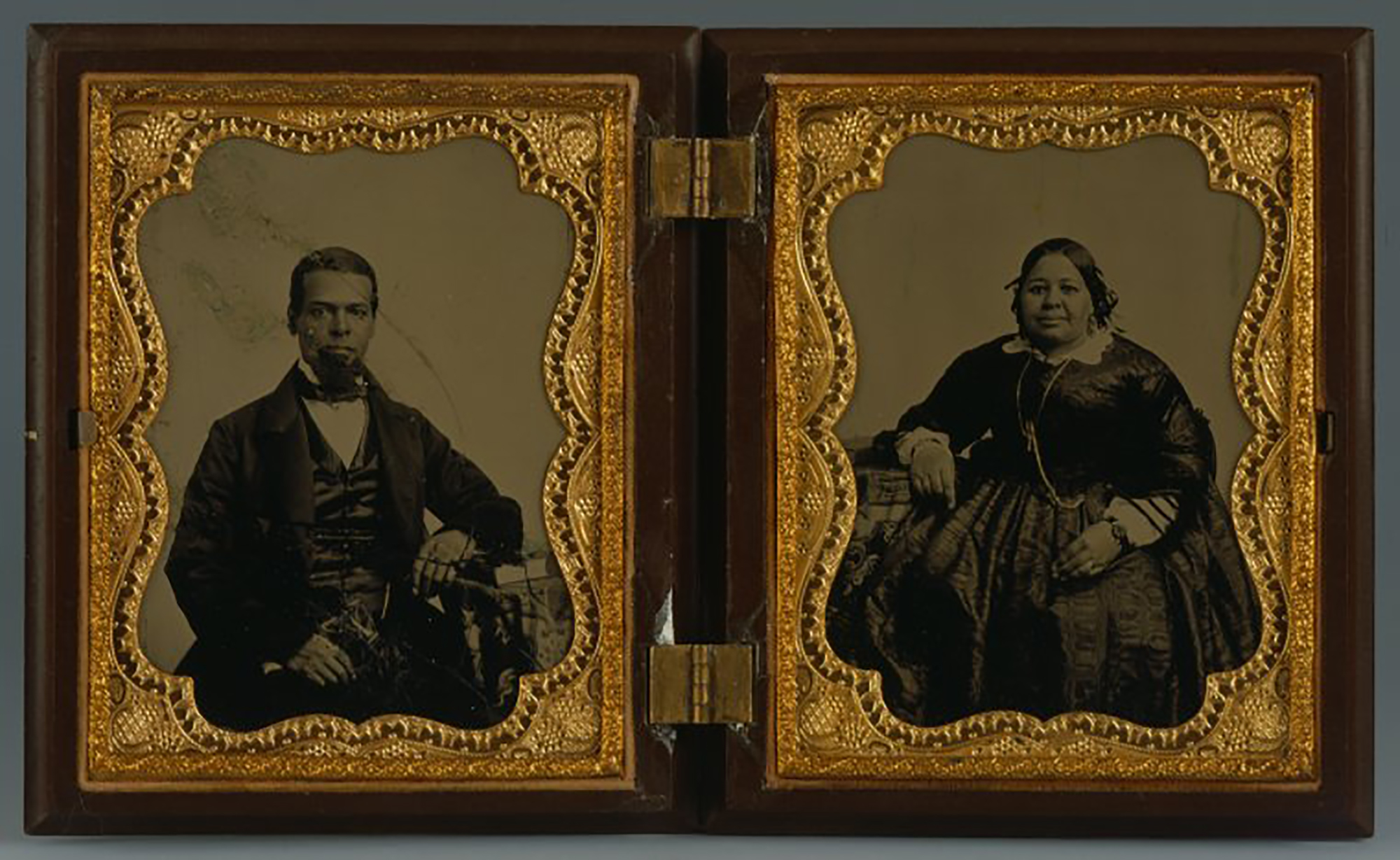 Deux photographies en noir et blanc dans un coffret bois et or à charnière. L'homme, à gauche, et la femme, à droite, sont noirs et portent un costume et une robe sombres à la mode.