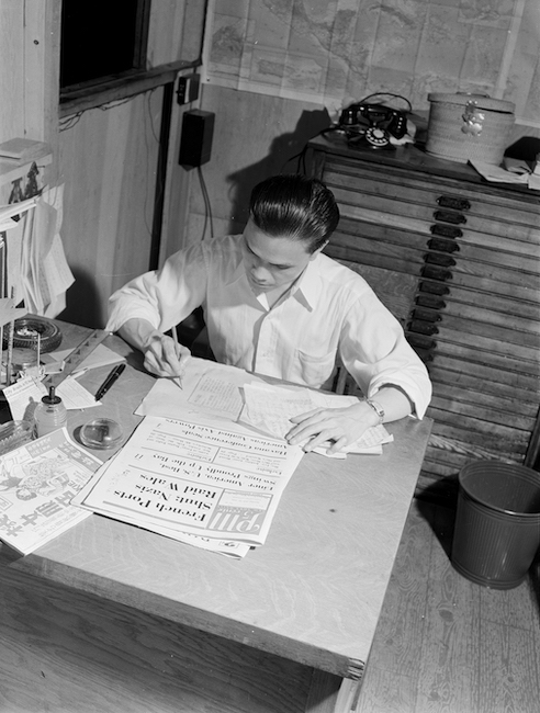 Photograph of a man sitting at a table surrounded by newspapers