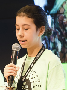 Une photo d'Anna Kathawala parlant avec un microphone. Elle porte un t-shirt vert et un cordon noir.