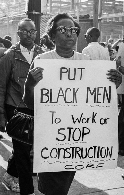 A protester at the 1963 CORE picket of the Downstate hospital construction site marches while holding a sign that reads "Put black men to work or stop construction."