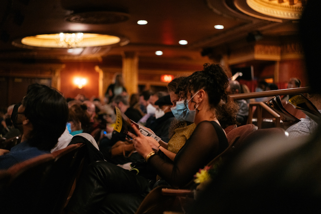 Photographie montrant l'intérieur d'un théâtre avec des invités assis dans le public. Les gens sont masqués et regardent des affiches, les lumières de la maison sont allumées.