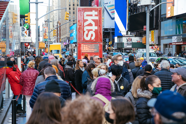 Photograph of crowds walking around Times Square, the tkts kiosk is visible in the center of the image.