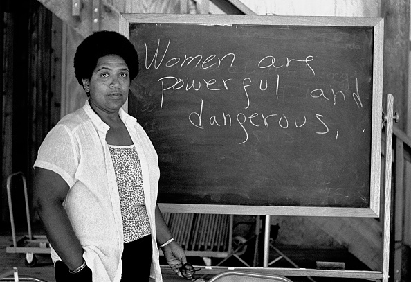 Une photo en noir et blanc d’une femme noire regardant fixement l’appareil photo. L'enseignement d'Audre Lorde, debout à côté d'un tableau noir sur lequel on peut lire "Les femmes sont puissantes et dangereuses".