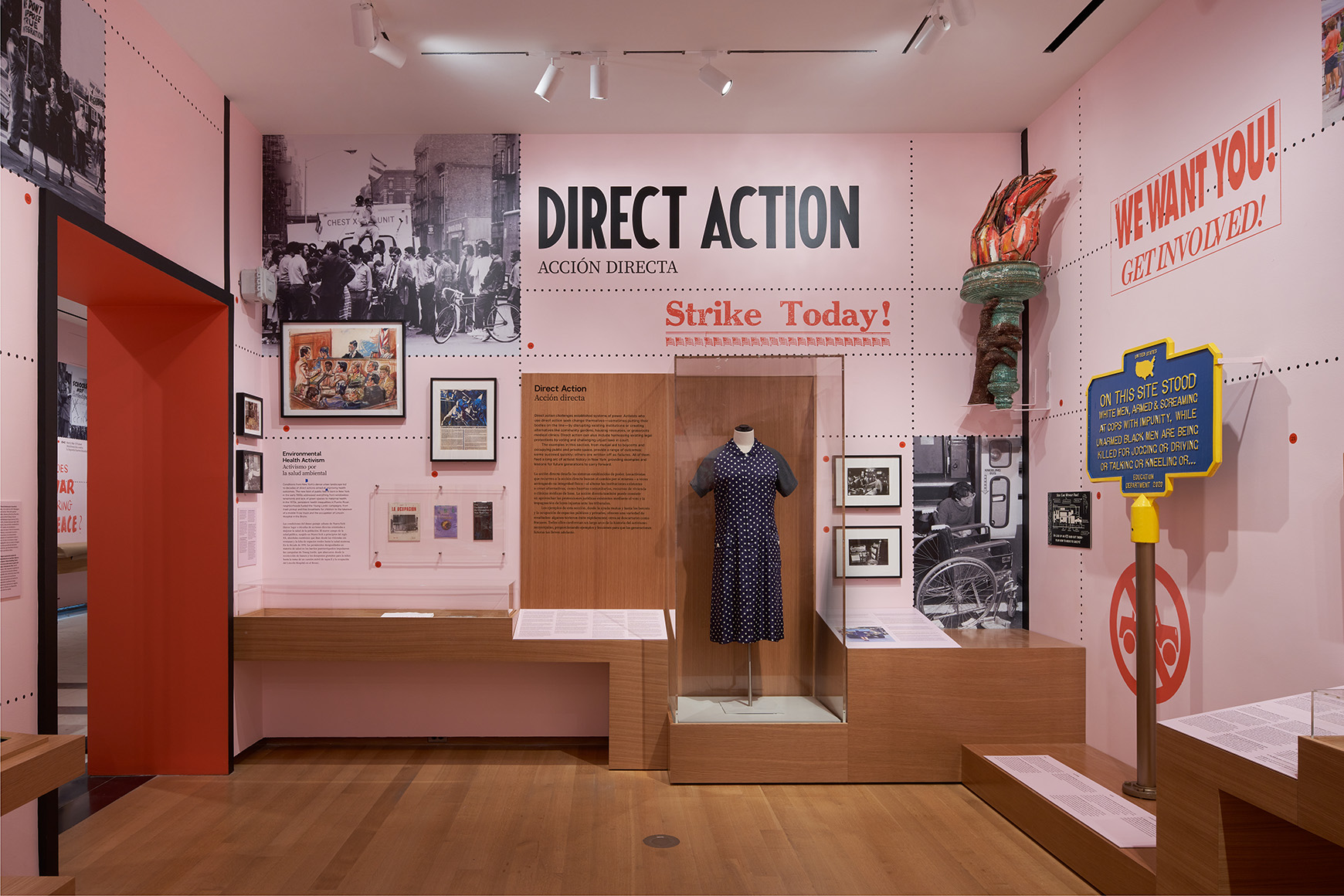 A gallery with pink walls and wooden cases showing photographs, sculptures, paintings, and clothing