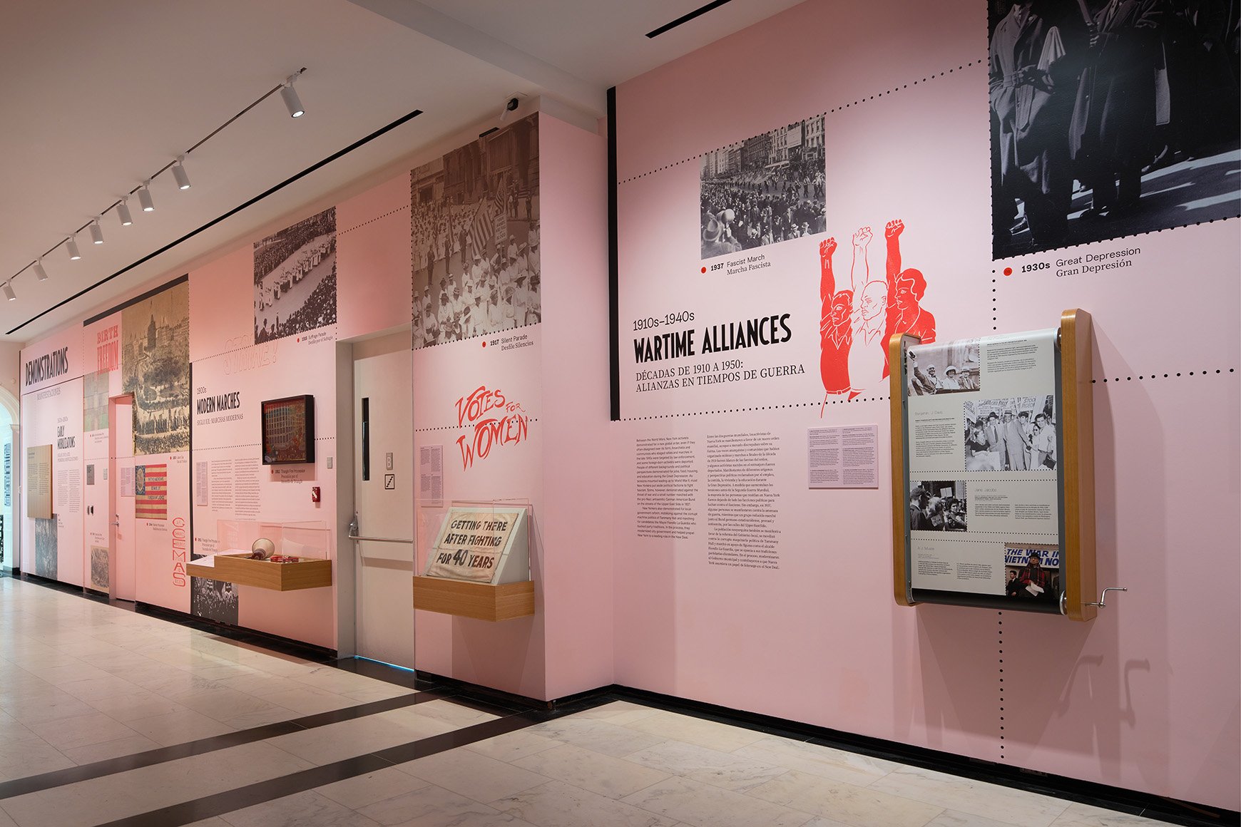A gallery with pink walls and wooden cases showing photographs, sculptures, paintings, and interactive displays