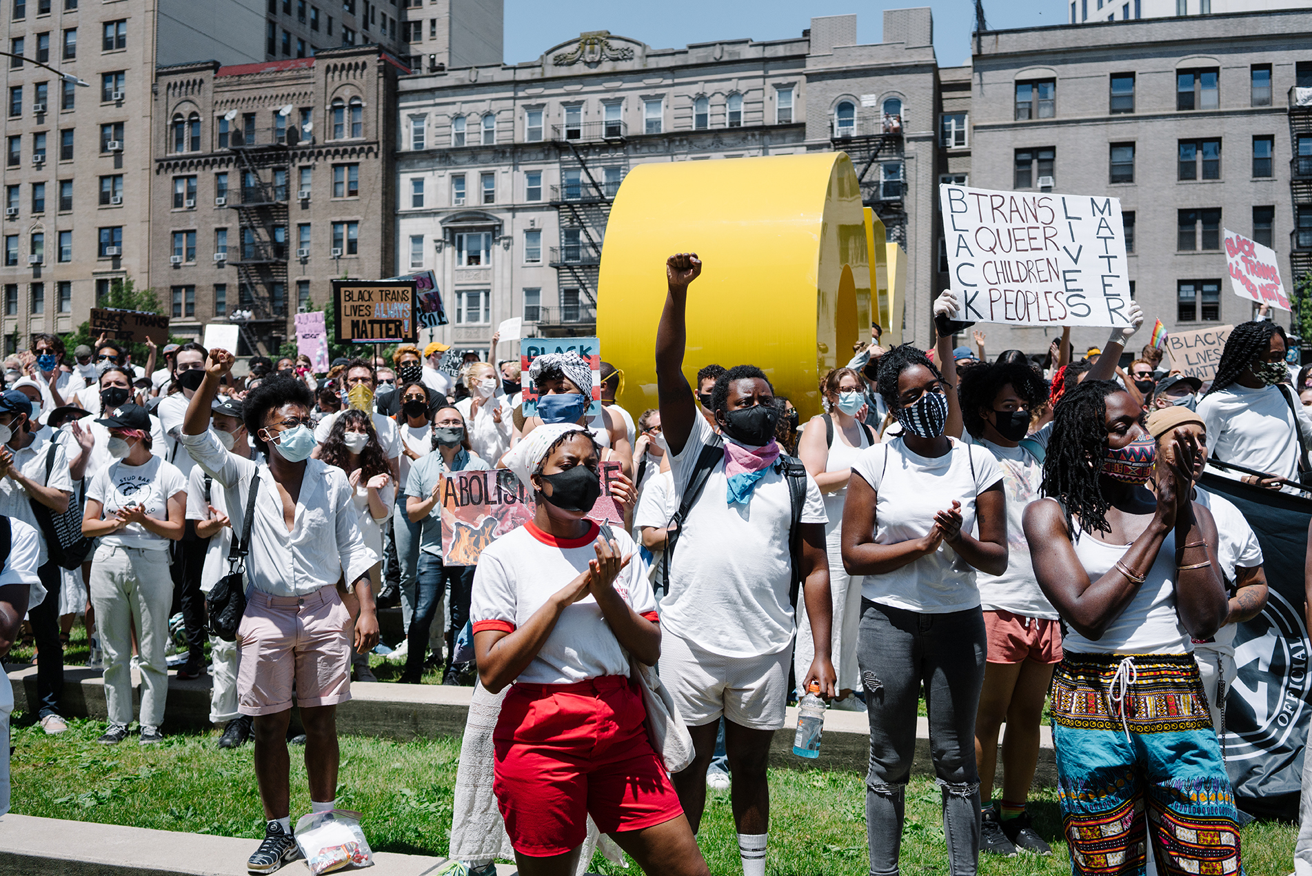 一群混血但主要是黑人的抗议者团结在布鲁克林博物馆外的广场上。 有的举着牌子，有的举着拳头。 都穿着白色。
