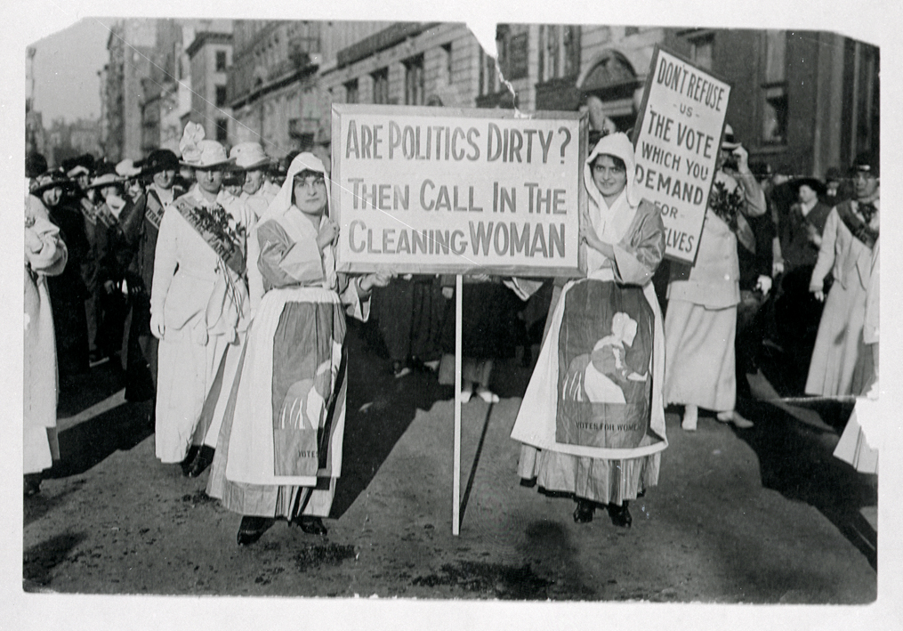 During a suffrage rally two women face the camera with a sign that says “Are politics dirty? Then call in the cleaning woman”
