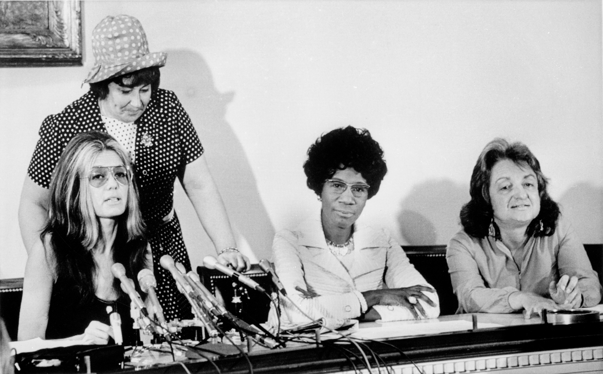 Three women sit at a table, one has microphones in front of her, suggesting it is a press conference 