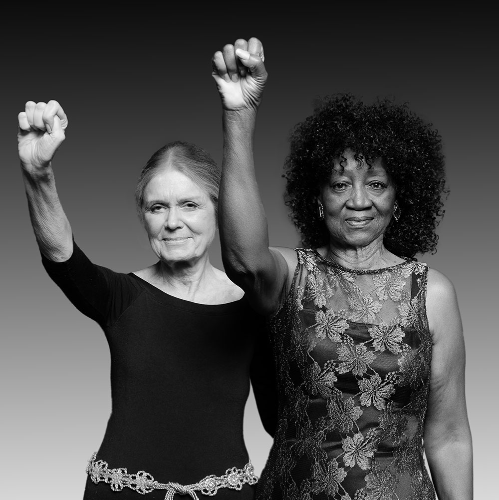 Duas mulheres mais velhas estão de frente para a câmera, cada um dos braços direitos erguidos em punho