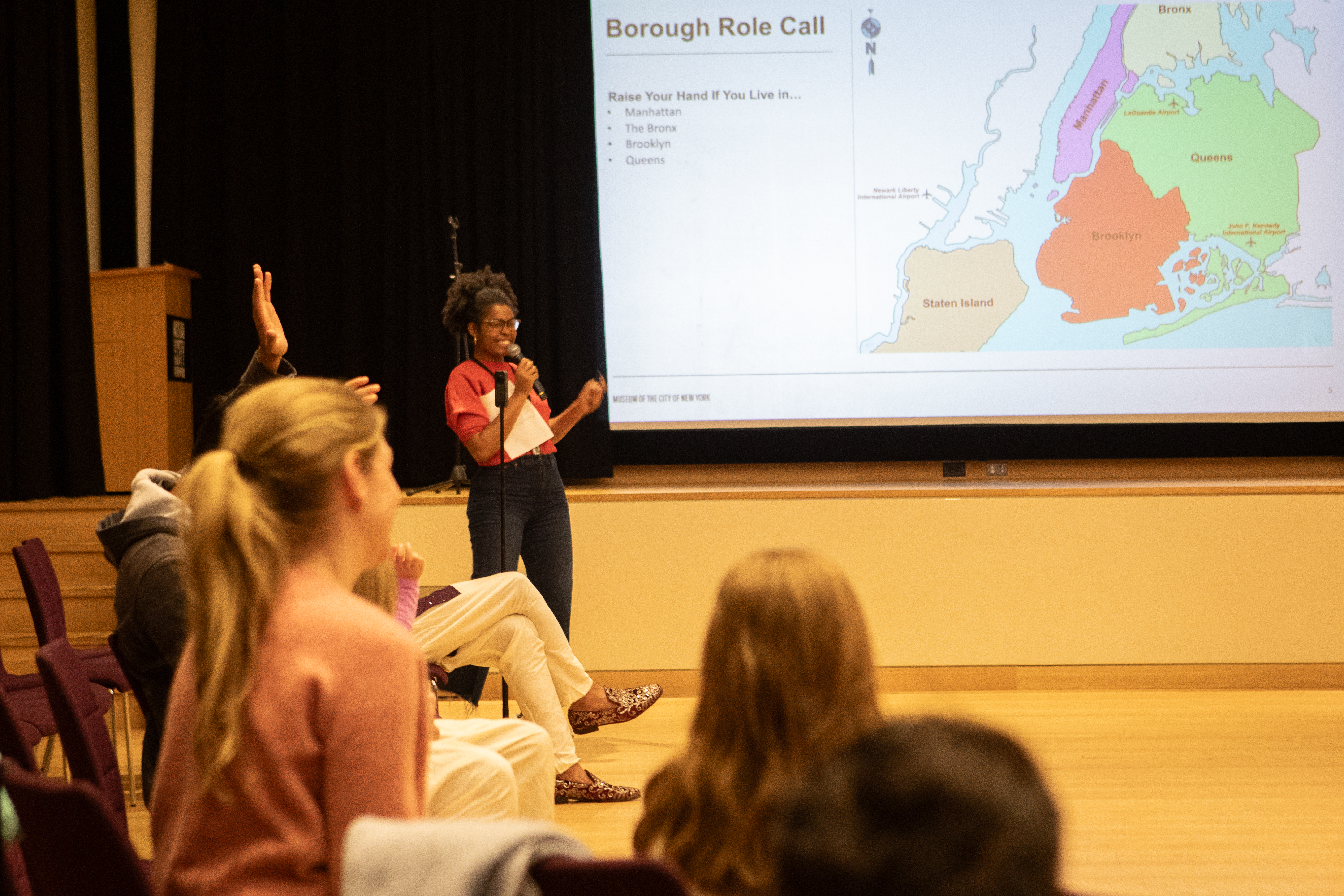 An image of someone smiling in front a slide that reads Borough Role Call with people participating with arms raised