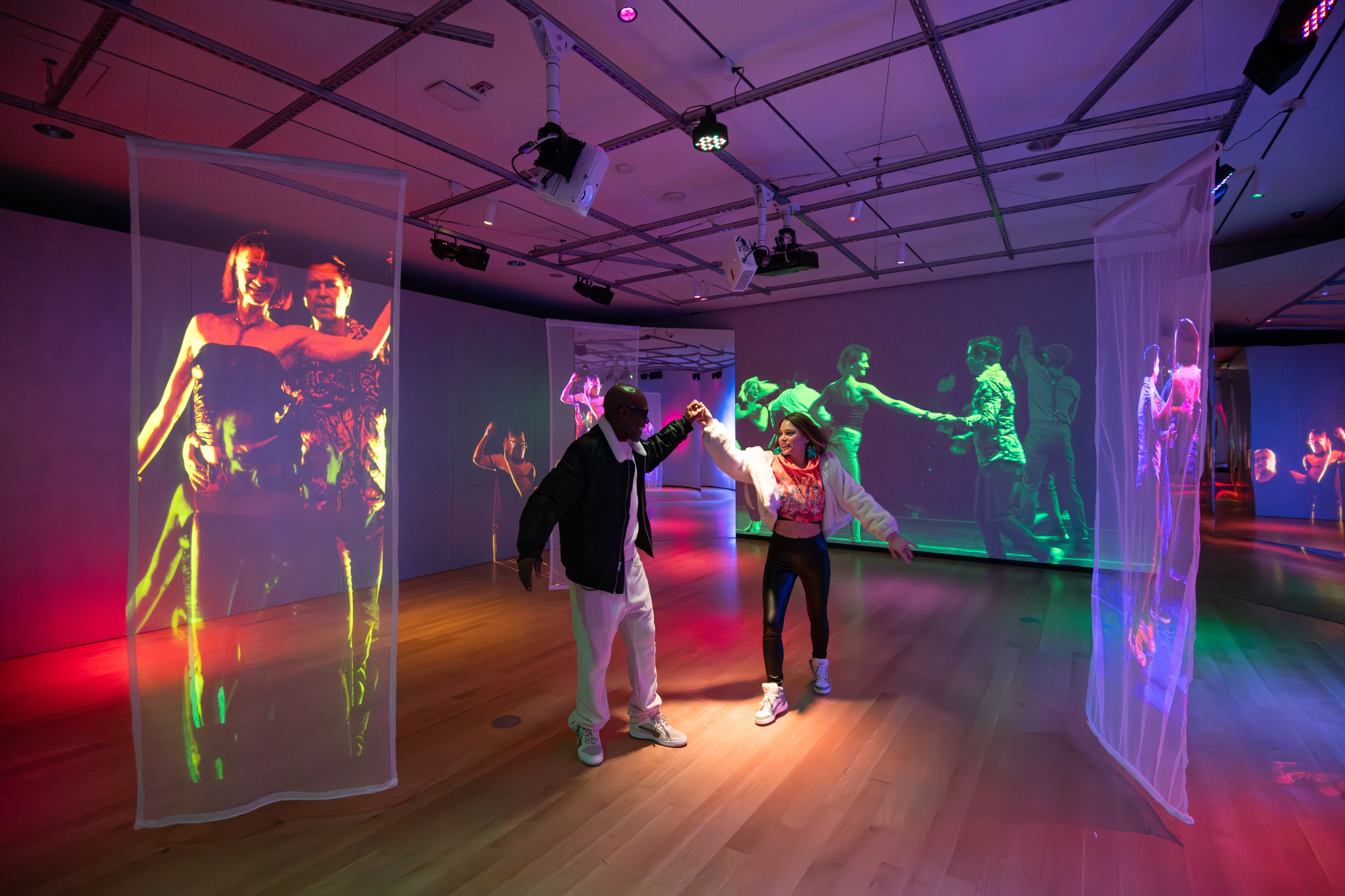 Two people dance together in a colorful, immersive art installation. The room features wooden floors and sheer fabric panels hanging from the ceiling, onto which vibrant, life-size video projections of dancers are displayed. The walls also show large-scale projections of people dancing, creating a lively, energetic atmosphere. Overhead lighting adds to the vivid, multicolored ambiance of the space.