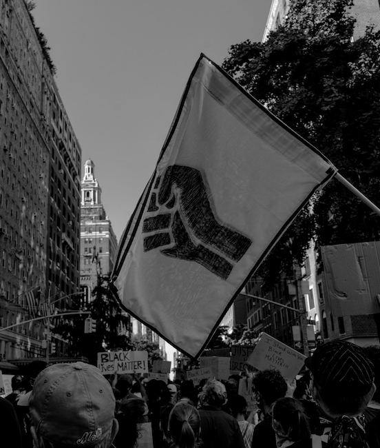 Black and white photo of a protest on NYC streets. In the center of the image is a flag with a black fist stencil.