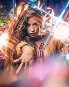 Headshot of Charlene Incarnate with long wavy light brown hair, a tan jacket, and posing with one hand out toward the camera.