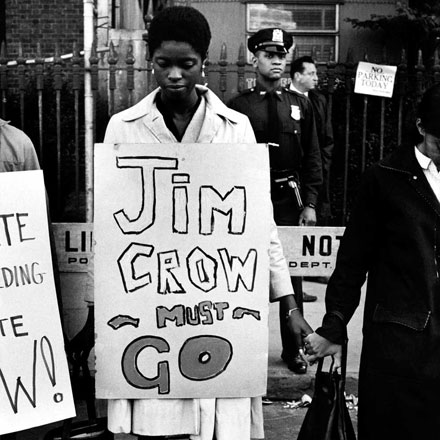 Una mujer se para en una fila de manifestantes tomados de la mano, con un cartel a su alrededor que dice "Jim Crow debe irse"