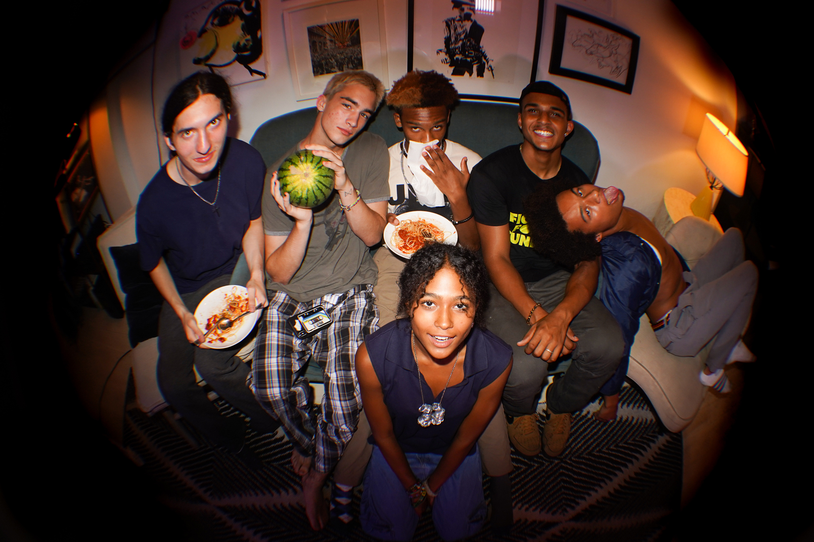 Six teenagers casually sitting on a couch. To the left, two of them are holding a slice a pizza and one is holding a watermelon. All of the teenagers are smiling. 