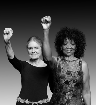 Gloria Steinem and Dorothy Pitman Hughes stand next to each other, each raising one arm with a closed fist.
