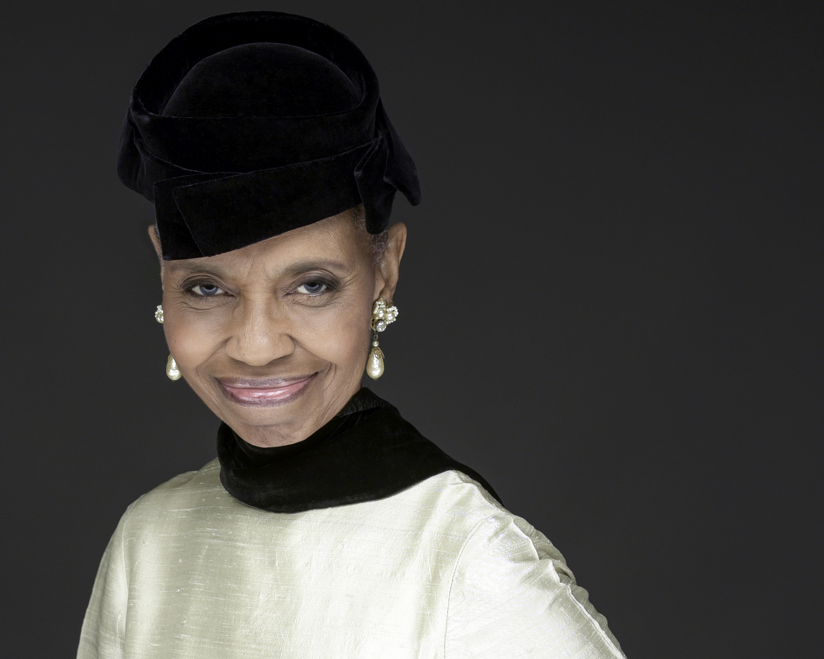 A portrait of an elegantly dressed Black woman smiling with closed teeth and looking directly at the camera