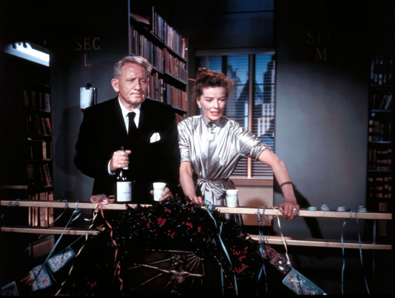 Spencer Tracy and Katharine Hepburn are standing next to each other and leaned on a rail. They are looking toward the lower right-hand corner of the screen. 