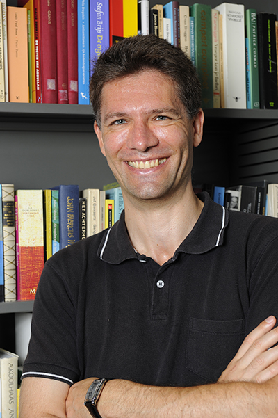 Headshot of a smiling man wearing a black, collared shirt standing arms-crossed in front of a bookcase.