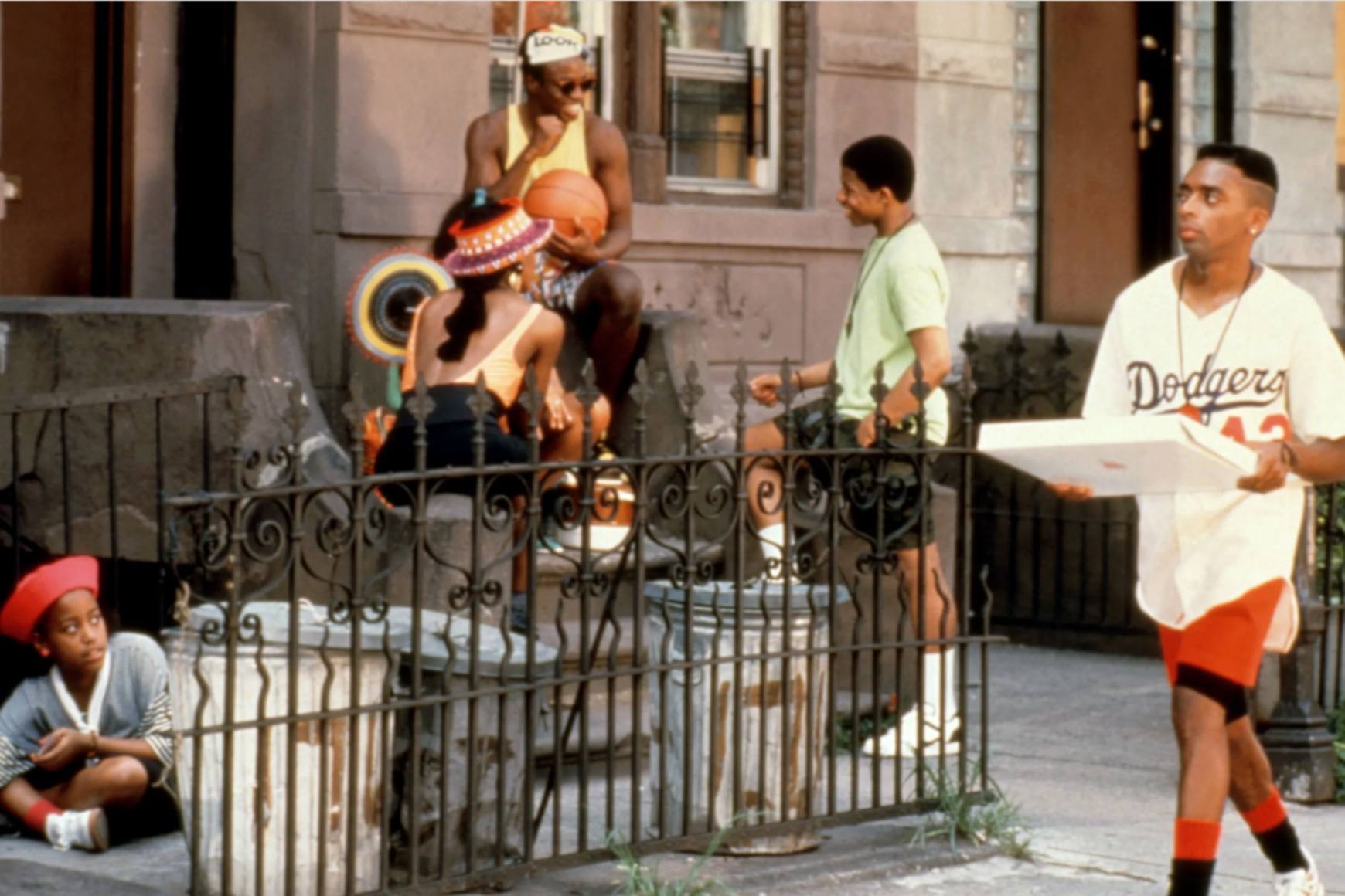斯派克·李 (Spike Lee) 饰演穆基 (Mookie) 走在贝德福德-史岱文森 (Bedford-Stuyvesant) 的街道上，人们在门廊上闲逛。