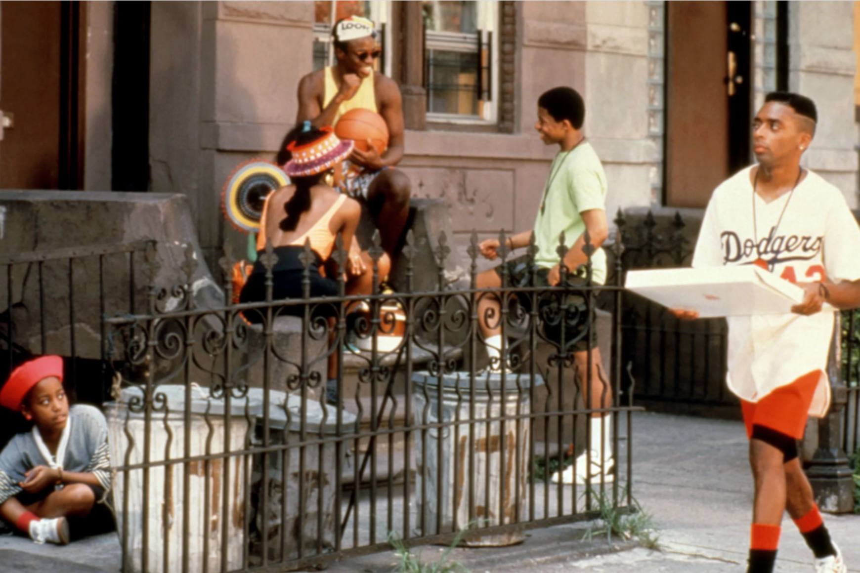 斯派克·李 (Spike Lee) 饰演穆基 (Mookie) 走在贝德福德-史岱文森 (Bedford-Stuyvesant) 的街道上,人们在门廊上闲逛。